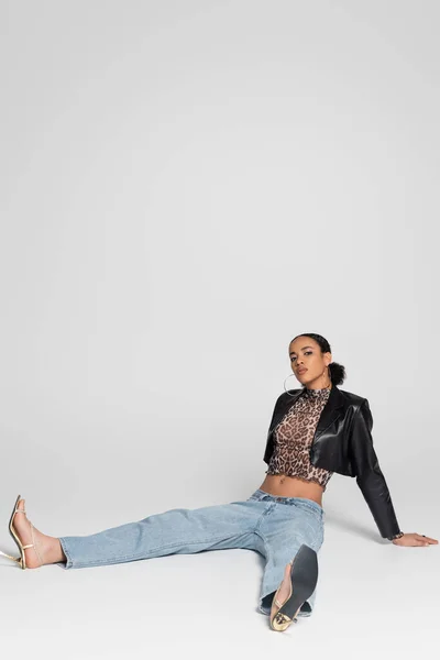 Piena lunghezza della giovane donna afroamericana in sandali con tacco alto e vestito alla moda seduta sul grigio — Foto stock