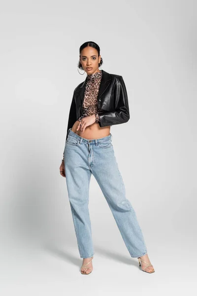 Piena lunghezza della donna afroamericana alla moda in sandali con tacco alto e jeans alla moda in piedi sul grigio — Foto stock