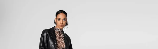 Стильна афроамериканська жінка в модній шкіряній куртці та сережках кільця ізольована на сірому, банер — стокове фото