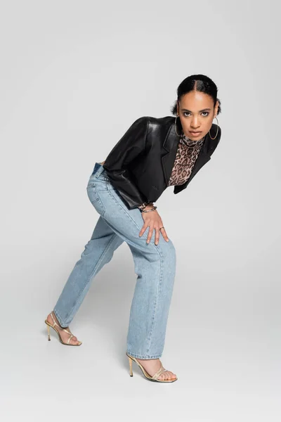 Pleine longueur de femme afro-américaine à la mode dans une veste à la mode et des sandales à talons hauts posant sur le gris — Photo de stock