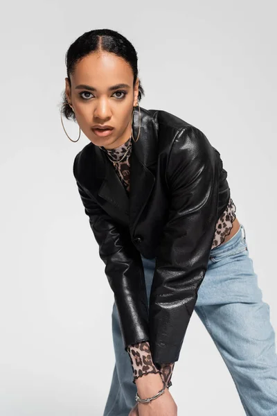 Mujer afroamericana de moda en chaqueta recortada de moda y vaqueros azules mirando a la cámara aislada en gris - foto de stock