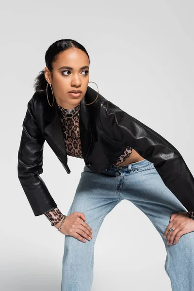 Fashionable african american woman in trendy cropped jacket and blue jeans looking away while posing isolated on grey — Stock Photo