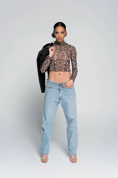 Volle Länge der afrikanisch-amerikanischen Modell in Crop Top mit Animal Print hält schwarze Jacke und steht mit der Hand in der Tasche auf grau — Stockfoto