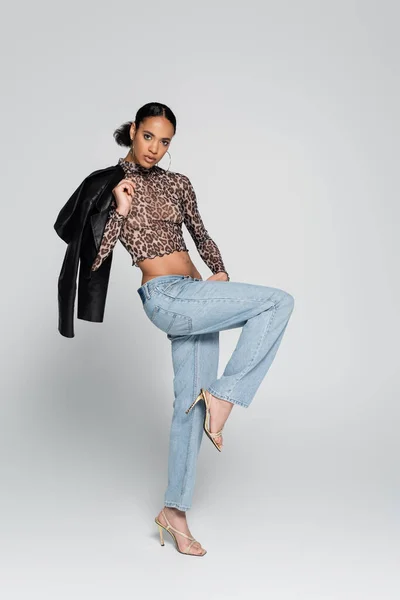 In voller Länge modische afrikanisch-amerikanische Modell in Crop Top mit Animal Print hält schwarze Jacke und posiert auf einem Bein auf grau — Stockfoto