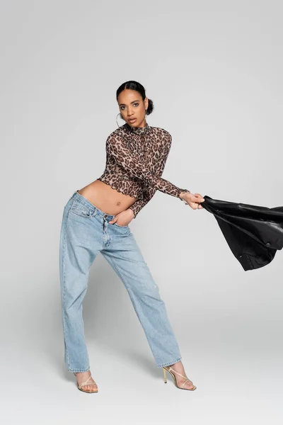 Full length of fashionable african american model in crop top with animal print holding black jacket while posing on grey — Stock Photo