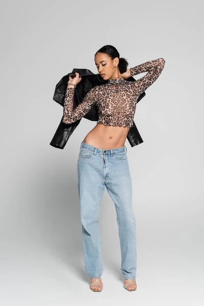 Full length of young african american model in crop top with animal print holding black jacket on grey — Stock Photo