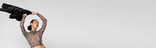 Modèle afro-américain à la mode en haut de culture avec imprimé animal tenant veste noire au-dessus de la tête isolé sur gris, bannière — Photo de stock