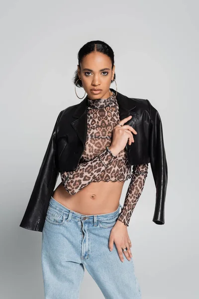 Stylish african american model in crop top with animal print wearing black leather jacket isolated on grey — Stock Photo