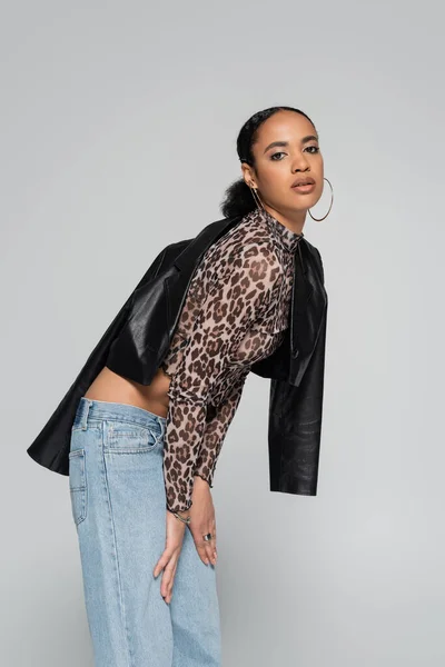 Mujer afroamericana con estilo en top de cultivo con estampado animal y chaqueta de cuero negro posando aislado en gris - foto de stock