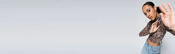Elegante modello afroamericano in crop top con stampa animalier e orecchini a cerchio gesticolando isolato su grigio, banner — Foto stock