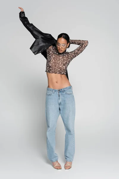 Full length of stylish african american woman in crop top with animal print and sunglasses wearing leather jacket on grey — Stock Photo