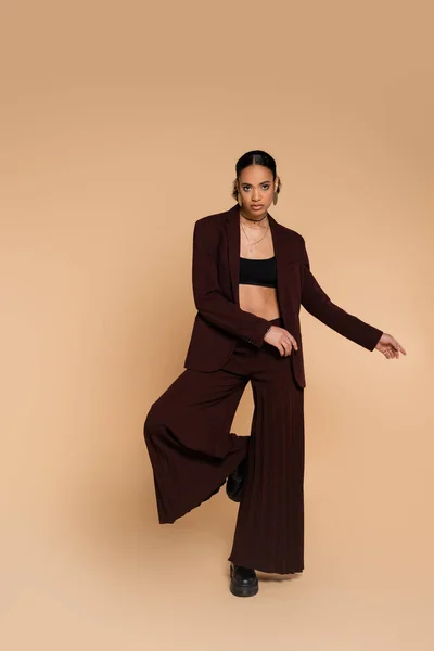 Full length of young african american woman in stylish suit with wide pants standing on one leg while posing on beige — Stock Photo
