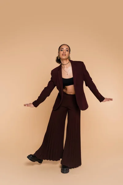 Full length of young african american woman in stylish suit with wide pants gesturing while posing on beige — Stock Photo