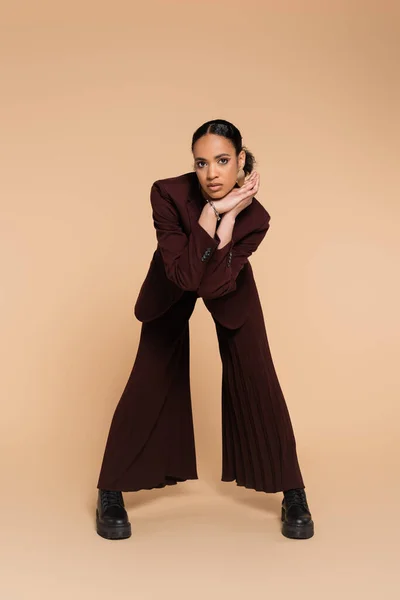 In voller Länge junge afrikanisch-amerikanische Frau in stylischem Blazer und kastanienbraunen weiten Hosen posiert auf beige — Stockfoto