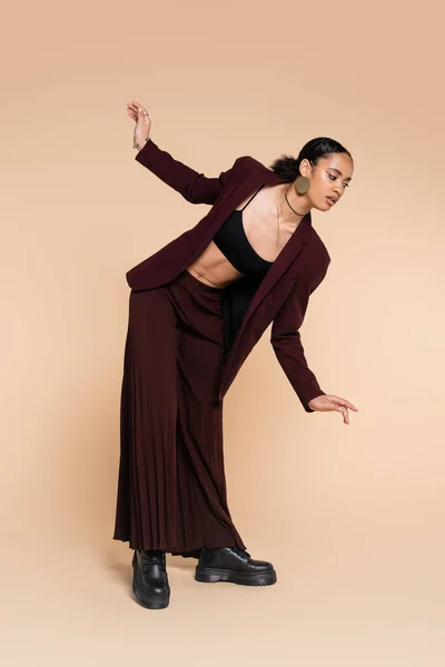 Comprimento total de mulher americana africana encantadora em terno maroon elegante com calças largas posando no bege — Fotografia de Stock