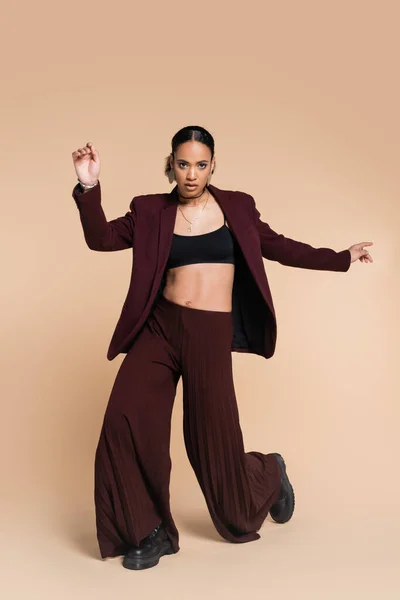 Comprimento total do jovem modelo afro-americano em terno marrom com calças largas posando em bege — Fotografia de Stock