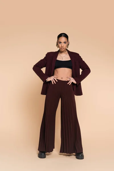 Full length of young african american woman in maroon suit with wide leg pants posing with hands on hips on beige — Stock Photo