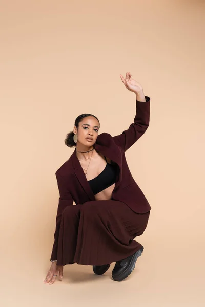 Longitud completa de la joven afroamericana en traje marrón posando con la mano levantada en beige - foto de stock