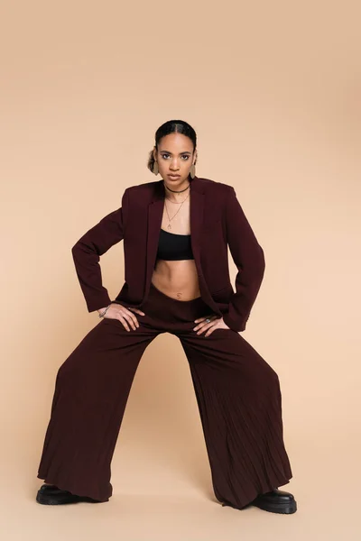 Full length of young african american woman in maroon suit with wide leg pants posing on beige — Stock Photo