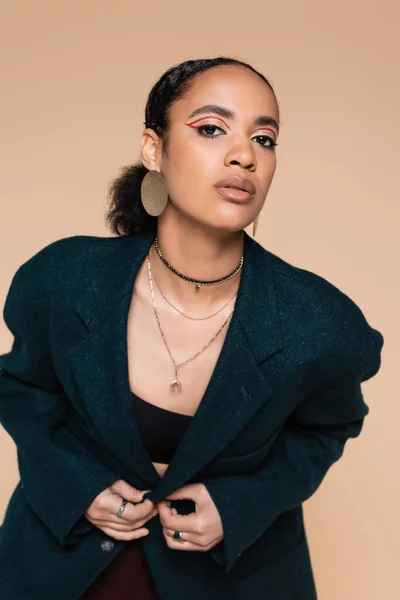 Joven modelo afroamericano con maquillaje posando en blazer de lana azul oscuro aislado en beige - foto de stock