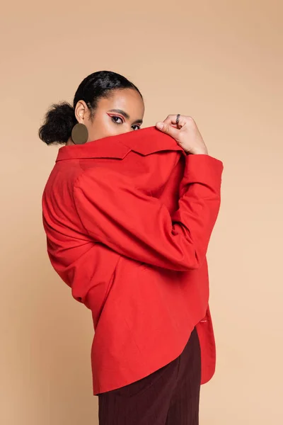 Portrait de brune modèle afro-américain en blazer rouge vif couvrant le visage tout en posant isolé sur beige — Photo de stock