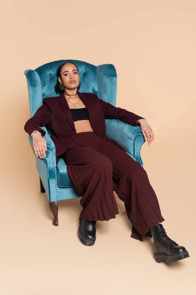 Stylish african american woman in maroon suit resting on blue velvet armchair on beige — Stock Photo