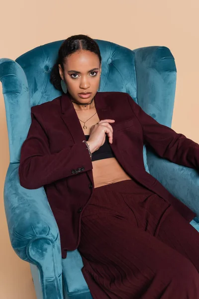 Stylish african american model in burgundy suit sitting on blue velvet armchair isolated on beige — Stock Photo