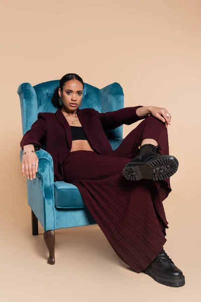 Stylish african american model in maroon blazer and wide pants sitting on blue velvet armchair on beige — Stock Photo