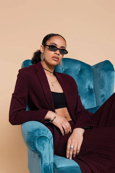 Stylish african american model in suit and trendy sunglasses sitting on blue velvet armchair isolated on beige — Stock Photo