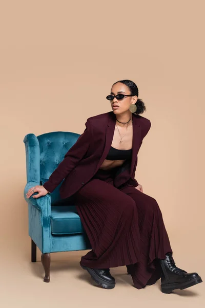 Modèle afro-américain pleine longueur en costume marron et lunettes de soleil tendance assis sur un fauteuil en velours bleu sur beige — Photo de stock