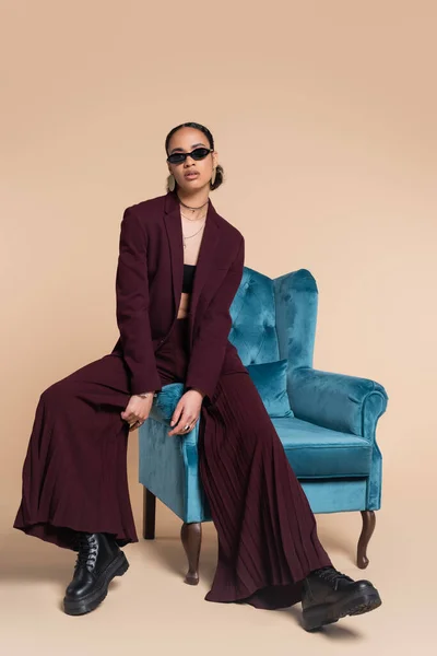 Comprimento total da mulher americana africana elegante em terno de borgonha e óculos de sol na moda sentado na poltrona de veludo azul no bege — Fotografia de Stock