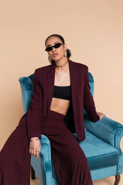 African american model in burgundy suit and stylish sunglasses sitting on blue velvet armchair on beige — Stock Photo