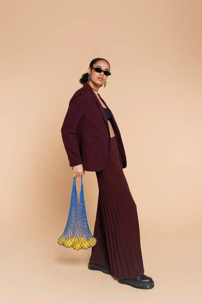 Full length of stylish african american woman in burgundy suit and sunglasses holding net bag with lemons on beige — Stock Photo