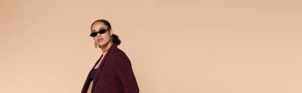 Стильная африканская американка в бордовом пиджаке и солнцезащитных очках, смотрящая на камеру, изолированную на бежевом, баннере — стоковое фото