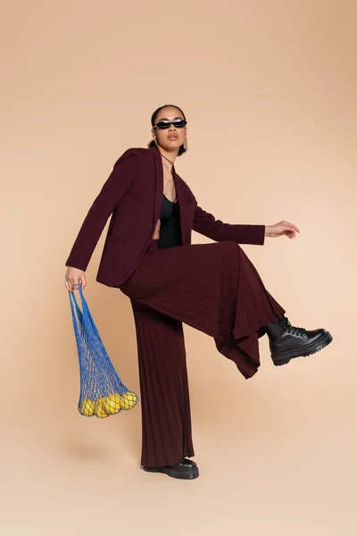 Pleine longueur de femme afro-américaine élégante en costume bordeaux et lunettes de soleil tenant sac en filet avec des citrons sur beige — Photo de stock