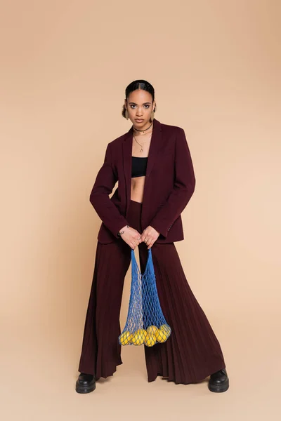 Full length of stylish african american woman in maroon suit holding mesh bag with lemons on beige — Stock Photo