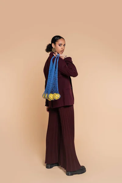 Longitud completa de mujer afroamericana bonita en traje marrón sosteniendo bolsa de malla con limones en beige - foto de stock