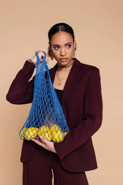 Stylish african american woman in burgundy suit holding mesh bag with lemons isolated on beige — Stock Photo