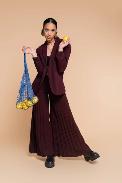 Full length of stylish african american model in burgundy wide pants and jacket holding mesh bag with lemons on beige — Stock Photo