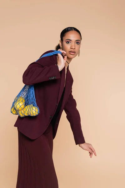 Élégante femme afro-américaine en veston bordeaux tenant sac filet avec des citrons frais isolés sur beige — Photo de stock