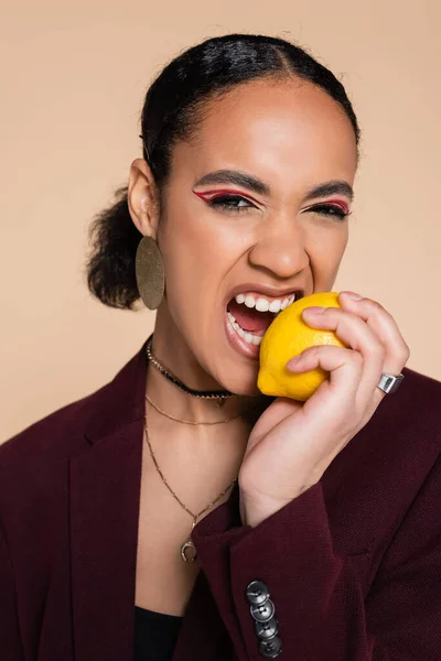 Afroamerikanerin im weinroten Blazer beißt in reife Zitrone auf Beige — Stockfoto