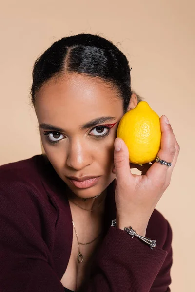 African american woman in burgundy blazer holding fresh lemon isolated on beige — Stock Photo