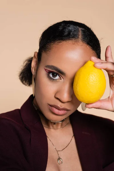 Африканская американка в бордовом пиджаке держит спелый лимон возле глаза, изолированного от бежевого — стоковое фото