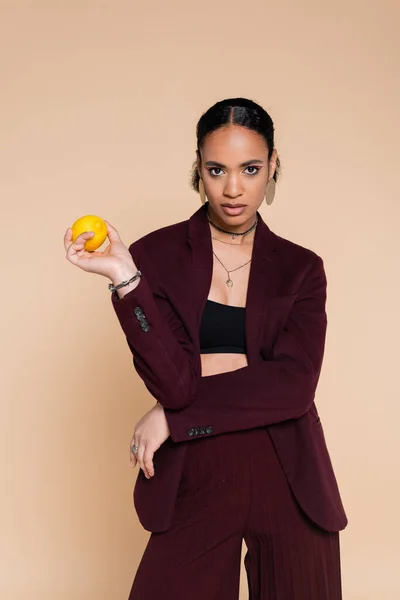 Afroamerikanerin in weinrotem Blazer posiert mit frischer Zitrone auf Beige — Stockfoto