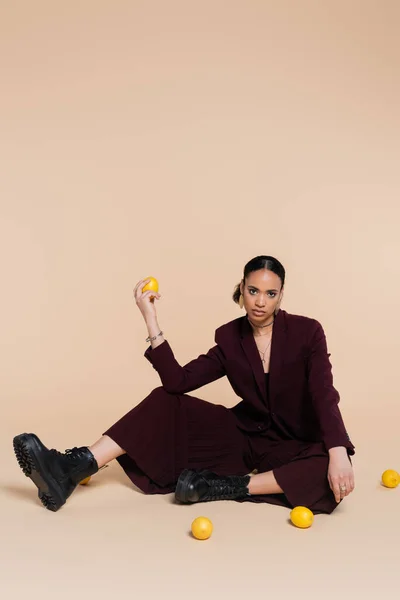 Modelo afroamericano con estilo en traje marrón sentado alrededor de limones frescos en beige - foto de stock