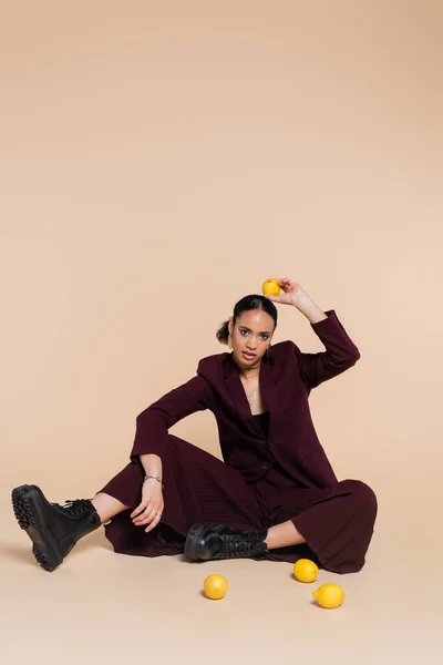 Young african american woman in stylish burgundy suit sitting around fresh lemons on beige — Stock Photo