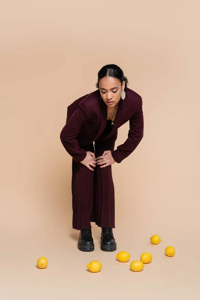 Stylische afrikanisch-amerikanische Frau im weinroten Anzug steht und auf frische Zitronen auf Beige blickt — Stockfoto