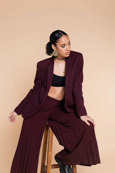 Brunette african american woman in stylish burgundy suit sitting on wooden high stool while posing isolated on beige — Stock Photo