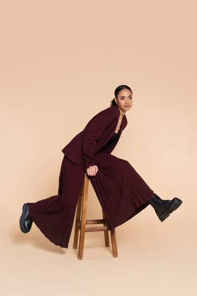 Comprimento total de mulher americana africana elegante em terno de borgonha posando em banquinho alto de madeira em bege — Fotografia de Stock