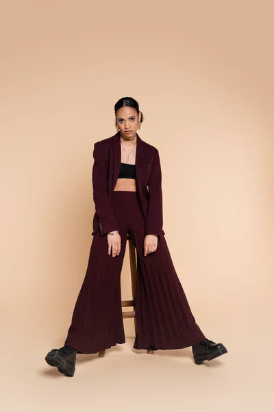 Full length of stylish african american woman in burgundy suit with wide pants standing near wooden high chair on beige — Stock Photo